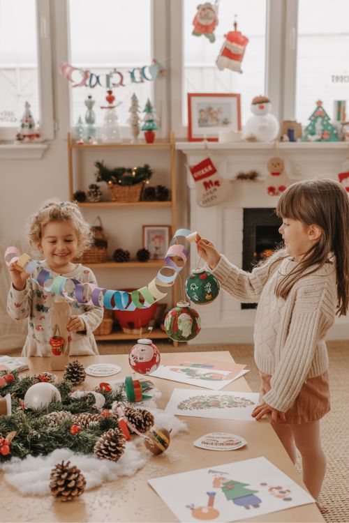 Living room decorated with kids’ DIY Christmas crafts, including paper chains, painted ornaments, garlands, pinecones, and faux snow, showing a joyful and budget-friendly family holiday scene.
