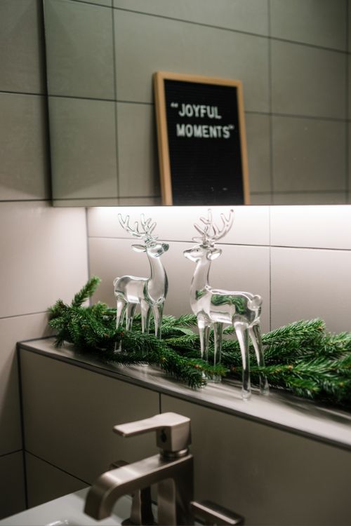Modern Christmas bathroom decor with glass reindeer figurines, floating shelf garland, and a holiday letter board.