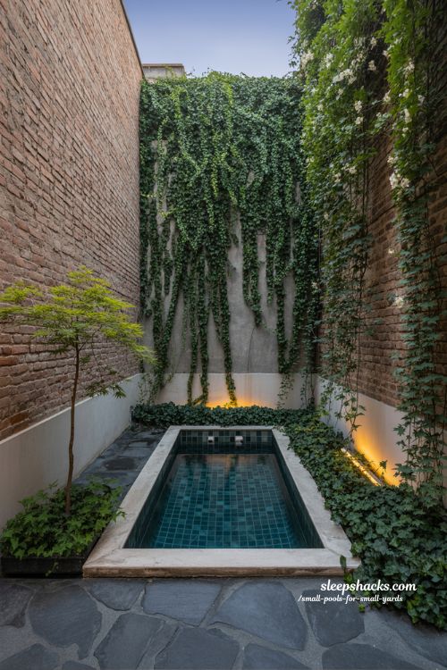 A candid, documentary-style photograph capturing a secluded, narrow plunge pool nestled deep within a very tight, private urban courtyard between two tall, weathered brick walls, radiating an intimate, "secret garden" feel. The pool is a simple, long rectangle with dark teal mosaic tiles and a very narrow polished concrete coping. To emphasize the vertical design, the brick walls stretch high up, almost completely enclosing the space, and are heavily draped in lush, cascading vines, ivy, and fragrant star jasmine that spill down toward the water. A slender, elegant Japanese maple tree is planted in a deep void in the concrete, its delicate leaves adding softness. The ground is made of large, irregular grey flagstones. Integrated warm, amber LED strip lighting is subtly hidden beneath the pool coping and among the base of the vines, casting a gentle, soothing glow that illuminates the water's surface and the textures of the brick and leaves. The setting is evening twilight, allowing the soft, inviting pool lights to become the focal point. A single, rich woven cotton throw blanket sits on a minimal wooden stool at the pool's edge, alongside a ceramic mug. The entire scene, from the wet flagstones in the foreground to the furthest leaf high on the wall, is in sharp, crystal-clear focus, capturing the intimate, realistic atmosphere with no blur. The natural evening light mixes beautifully with the warm, inviting artificial light.