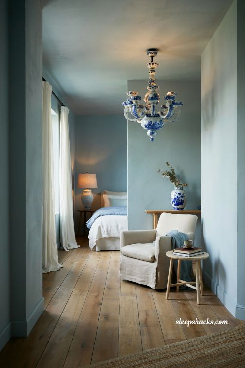 Eye-level wide-angle interior photography shot from a slightly off-center corner perspective capturing a serene coastal blue bedroom anchored by cerused oak nightstands with a whitewashed textured finish, with the camera framing the full bed area, bedside symmetry, and surrounding architectural balance in a clean editorial composition. The perspective emphasizes material texture, subtle reflection, and layered coastal softness while keeping every surface sharply defined and fully visible. The room reflects a calm coastal-inspired minimalist aesthetic where natural wood finishes are softened through a whitewashed cerused oak treatment, creating a light, breathable foundation that pairs effortlessly with blue bedding tones. The design feels relaxed, timeless, and quietly refined, like a well-curated beach house interior that evolves naturally over time. The color palette is composed of soft coastal blue, faded denim blue, pale aqua undertones, crisp white, warm ivory, cerused oak whitewashed wood, sandy beige, and driftwood gray, creating a balanced harmony between cool coastal tones and softened natural wood warmth. Key props and details include a bed dressed in crisp white linen bedding layered with soft coastal blue accents, creating a clean and airy focal point. On either side of the bed sit cerused oak nightstands featuring a lightly textured whitewashed finish with visible wood grain and subtle lime-washed effect. The nightstands include glass-front drawers that softly reflect ambient light, adding depth and a gentle sense of transparency to the bedside styling. Each surface is styled minimally with a ceramic table lamp, a small stack of books, or a single glass or porcelain vase in muted coastal tones. The furniture palette avoids heaviness, maintaining a light visual footprint that supports the overall airy coastal theme. Walls are warm white or off-white to enhance brightness and allow the cerused oak texture and blue bedding to stand out naturally. Flooring is light natural wood or driftwood-inspired planks that subtly echo the organic grain of the nightstands without overpowering them. Lighting is soft natural daylight filtering gently through unseen windows, highlighting the textured finish of the cerused oak and creating subtle interplay between matte wood grain and soft reflections from glass drawer fronts. The light remains calm and diffused, enhancing warmth without reducing clarity. Mood and atmosphere translate into soft coastal elegance and layered natural texture, where cerused oak introduces quiet sophistication, blue textiles bring freshness, and whitewashed finishes maintain lightness and ease. The room feels balanced, airy, and timeless, like a coastal retreat designed for longevity and gentle evolution. Coastal blue bedroom ideas with pale blue linen curtains and airy light filled beach house style (1)