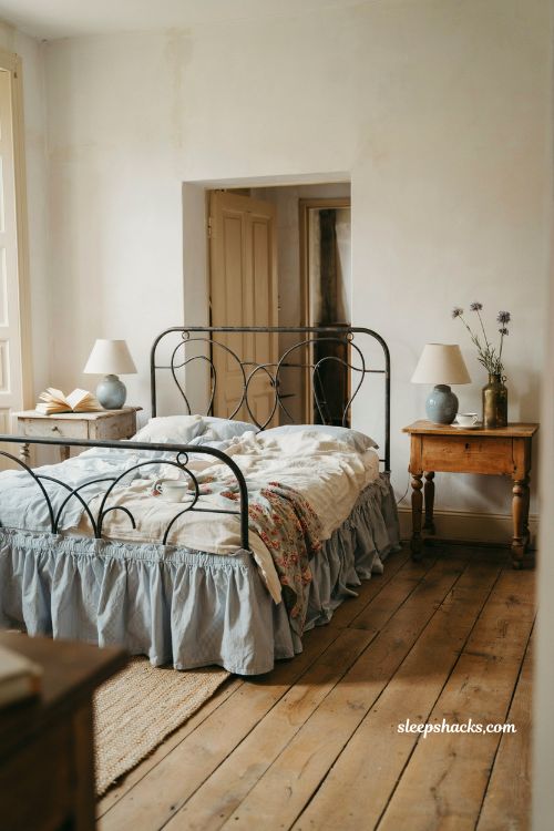 Eye-level wide-angle interior photography shot from a slightly off-center perspective capturing an elegant French-inspired bedroom designed around soft blue wallpaper, with the camera framing the full bed wall composition, side furnishings, and architectural balance in a clean editorial layout. The composition highlights texture, restraint, and hotel-like refinement while maintaining a warm, livable atmosphere. The room reflects a boutique French country aesthetic with subtle luxury influence, where pattern, fabric, and light work together to create a restful yet elevated environment. The design is calm and intentionally minimal, with careful layering of classic materials rather than decorative excess. The color palette is composed of soft powder blue, muted sky blue wallpaper tones, crisp white linen, warm ivory, aged oak brown, soft beige, and brushed brass gold accents, creating a balanced contrast between freshness and warmth. Key props and details include soft blue wallpaper covering the main feature wall, featuring a small-scale, understated pattern such as faint stripes, delicate floral motifs, or a subtle damask texture that reads more as surface depth than bold decoration. Centered against the wall is a bed with a gently upholstered headboard in a neutral fabric, adding softness and structure. The bedding is layered in crisp white linens with clean folds and subtle texture, creating a fresh boutique-hotel feel. On either side, simple nightstands in light wood or painted neutral tones maintain visual lightness, styled with restraint—each holding a brass-accented lamp or vintage-inspired lighting piece with warm metallic detailing. A decorative brass mirror or antique-style wall accent may be placed subtly to reflect light and add historical character without overwhelming the composition. The floor is warm natural wood, grounding the cooler blue tones and adding tactile warmth. Lighting is soft natural daylight diffused through large unseen windows, evenly illuminating the wallpaper so its delicate pattern remains visible without overpowering the space. The light enhances the crispness of white linens while gently warming brass and wood elements for balance. Mood and atmosphere translate into quiet sophistication and restful clarity, where soft blue wallpaper introduces gentle texture, white linens create purity and freshness, and vintage accents add understated character. The room feels polished yet calm, like a serene boutique hotel suite designed for comfort, balance, and timeless appeal. French blue bedroom ideas with blue and white toile bedding and old world cottage charm (3)