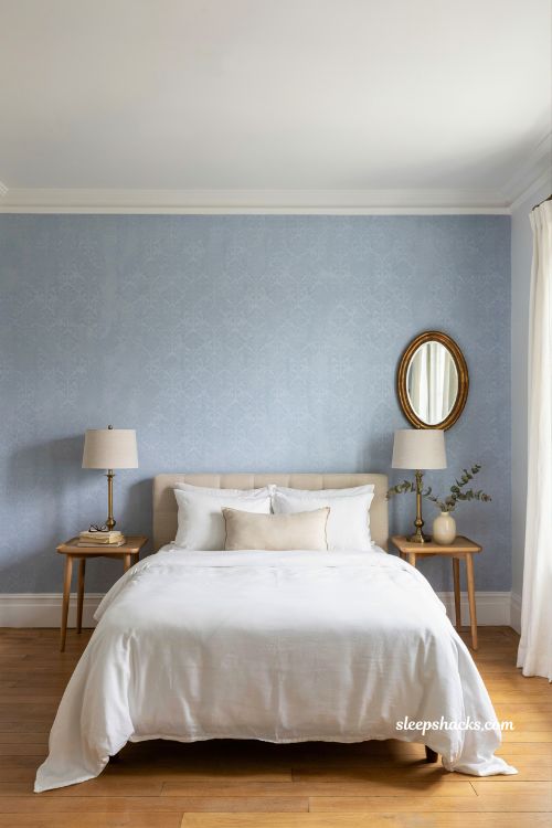 Eye-level wide-angle interior photography shot from a slightly angled corner perspective capturing a refined French-inspired bedroom where soft blue walls form the dominant architectural backdrop and warm yellow and gold accents are used as intentional visual highlights, with the full bed composition, side furnishings, and surrounding space clearly visible in a balanced editorial layout. The framing emphasizes color contrast and restrained styling while keeping the room airy and cohesive. The room reflects understated French country elegance with a subtle contemporary lift, where a cool base palette is gently energized through carefully placed warm accents. The design feels thoughtful, balanced, and quietly expressive rather than decorative or busy. The color palette is composed of dusty French blue, muted sky blue undertones, warm ivory, flax beige, aged oak brown, soft mustard yellow, and brushed gold tones, creating a harmonious interplay between cool serenity and warm vibrancy. Key props and details include a bed centered against soft blue walls, dressed in neutral linen bedding in warm ivory or flax tones to maintain calm visual grounding. Accent elements introduce warm yellow and gold details in a restrained, purposeful way—such as a single mustard-striped throw pillow placed at the front of the bed, a small golden table lamp on a bedside table, or a subtle framed artwork featuring soft yellow-toned abstract or botanical forms. These accents are carefully spaced so they read as highlights rather than dominant features. Bedside tables are simple and understated in light wood or neutral paint finishes, keeping focus on the color interplay. A warm brass or gold hardware detail appears on drawer pulls or lamp bases, reinforcing cohesion across accents. The flooring is aged natural wood, adding warmth and depth beneath the cooler blue environment. Lighting is soft natural daylight diffused through unseen windows, balanced with gentle warm reflections from gold accents that subtly enrich the blue tones rather than overpower them. The light remains even and calm, preserving clarity across textures and maintaining a breathable atmosphere. Mood and atmosphere translate into balanced contrast and quiet liveliness, where cool blue establishes calm structure, warm yellow introduces gentle energy, and gold accents add refinement and cohesion. The room feels elegant but not rigid, softly uplifted by color harmony that keeps the space feeling fresh, inviting, and emotionally warm. French blue bedroom ideas with blue and white toile bedding and old world cottage charm (4)