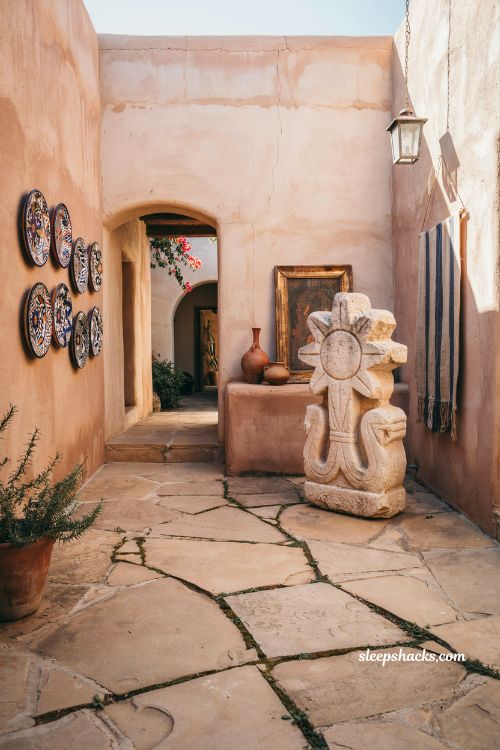 Eye-level wide-angle editorial interior photography shot of a Mexican hacienda patio fire pit gathering corner, composed to emphasize intimacy, warmth, and social flow within a rustic courtyard setting. The camera frames a circular seating arrangement around a central fire feature, showing how the space naturally draws people inward even in an open-air environment. The space reflects traditional Mexican hacienda design blended with functional outdoor living, where adobe walls, terracotta flooring, and wrought-iron details form a grounded architectural envelope around a cozy fire-focused seating zone. Subtle hand-painted tile accents appear along nearby edges, tying the fire pit corner visually to the broader patio design language. The color palette is built from glowing ember orange, deep charcoal black, sun-baked terracotta, dusty adobe beige, warm sandstone, soft cream textiles, and muted earthy greens from surrounding plants, all gently enhanced by firelight and fading evening sky tones. Key props and details include a central fire pit or fire table—either a low stone basin or a sleek propane fire feature with controlled flame—surrounded by rustic yet comfortable seating arranged in a soft circular layout. The fire pit surround is crafted from stone, terracotta, or aged masonry to visually connect with the hacienda architecture. Weather-resistant cushions in neutral woven fabrics are placed on benches and lounge chairs, showing subtle texture and slight compression from use. A quick-dry outdoor rug anchors the seating area, adding softness underfoot while maintaining a practical outdoor materiality. Nearby terracotta planters and clay pots soften the edges of the zone, while wrought-iron elements and wooden accents subtly echo the broader patio structure. Lighting is dominated by the warm, flickering glow of the fire, casting dynamic highlights and soft moving shadows across faces of stone, fabric, and clay surfaces. As evening deepens, residual ambient light from the courtyard blends with the firelight, creating a layered warm atmosphere without harsh contrast. Mood and atmosphere translate into natural gathering energy, where the fire becomes an unplanned social anchor, aged textures amplify warmth and familiarity, and simple layered furnishings create a space that feels effortless, welcoming, and quietly magnetic. The corner feels like the emotional center of the patio, where conversations naturally extend and time slows down. no people, no text, no watermarks, no blur, no bokeh, all surfaces in sharp focus, photorealistic, 4K resolution, editorial interior photography, Pinterest-worthy