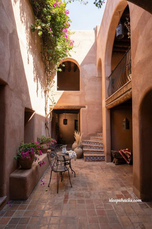 Aerial three-quarter wide-angle shot from eye level looking slightly down into a sun-warmed courtyard patio framed by adobe walls, capturing the full spatial composition of an intimate Mexican hacienda outdoor living space. A rustic central seating area is arranged around a low stone coffee table, surrounded by textured stucco walls in warm terracotta and faded adobe tones that show natural age and subtle imperfections. The space reflects traditional Mexican hacienda design blended with artisan craftsmanship and timeless architectural heritage. The color palette is deeply grounded in sunbaked terracotta, clay red, sandy beige, muted ivory, weathered brown wood, and soft oxidized copper accents that gently reflect the fading golden light. Key props and details include hand-painted Talavera tile accents along stair risers and wall niches, wrought-iron railings and lantern holders with intricate traditional curves, aged wooden beams, clay pottery planters filled with drought-tolerant greenery, and multiple copper lanterns and planters placed thoughtfully near seating corners. The copper surfaces show natural patina, glowing softly where late afternoon sun touches them, becoming a focal point that subtly elevates the entire courtyard atmosphere. Lighting is warm, low-angle golden hour sunlight filtering across the courtyard, casting soft architectural shadows along textured walls and highlighting the imperfections in plaster and tile without harsh contrast. The light feels slow, lingering, and calm, as if the day is gently dissolving into evening. Mood and atmosphere emphasize soft earthy colors that naturally calm the eye, aged textures that give depth and authenticity, and simple handcrafted details that create a sense of lived-in warmth and cultural continuity. The space feels like a quiet retreat where time slows down, blending rustic heritage with understated elegance and comfort. Mexican hacienda patio ideas with reclaimed wood furniture rustic beams salvaged timber courtyard design weathered farmhouse outdoor space warm natural textures (1)