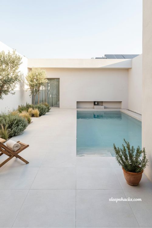 A wide-angle cinematic architectural photography shot from a slightly elevated poolside perspective, capturing a luxurious tropical villa swimming pool fully embraced by lush greenery. The composition emphasizes immersion and escape, where the pool feels like a hidden resort oasis surrounded by dense natural planting that blurs the boundary between architecture and landscape. The water becomes the calm centerpiece of a private jungle-inspired retreat designed for relaxation and sensory escape.The style and era reflect modern tropical resort villa design with biophilic and Balinese-inspired influences, focusing on harmony between natural materials, vegetation, and open-air living. The surrounding architecture is understated, featuring warm stucco walls, natural wood accents, and open transitional spaces that allow greenery and water to dominate the visual experience.The color palette consists of deep emerald and layered jungle greens, terracotta and clay-toned tiles, warm sandy beige stone, rich teak wood accents, and reflective turquoise-blue water. Palm shadows fall softly across the pool surface, creating dynamic natural patterns that shift with the movement of sunlight and leaves.Key props and details include a free-flowing rectangular or softly curved pool design, terracotta-toned or earthy matte tiles around the deck, dense tropical planting such as palm trees, banana leaves, ferns, and climbing vines, and a few minimalist lounge chairs positioned subtly within the greenery. The planting is intentionally layered and immersive but carefully maintained to preserve openness and cleanliness.Lighting is soft golden-hour natural sunlight filtering through palm fronds, casting patterned shadows across the water and stone surfaces. The light feels warm, dappled, and organic, enhancing texture and depth while reinforcing the natural resort-like atmosphere of the space.Mood and atmosphere evoke tropical serenity and vacation-like escape — rich natural greens that feel alive and immersive, warm earthy textures that ground the space, and gentle water reflections that create a calming rhythm throughout the environment. The entire setting feels like a private jungle villa retreat, peaceful, lush, and emotionally transportive.built in jacuzzi seating pool design with luxury marble look spa edge and modern villa relaxation aesthetic (1)