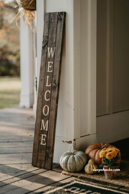 Rustic wooden welcome sign leaning against front porch door frame with fall pumpkins and mums