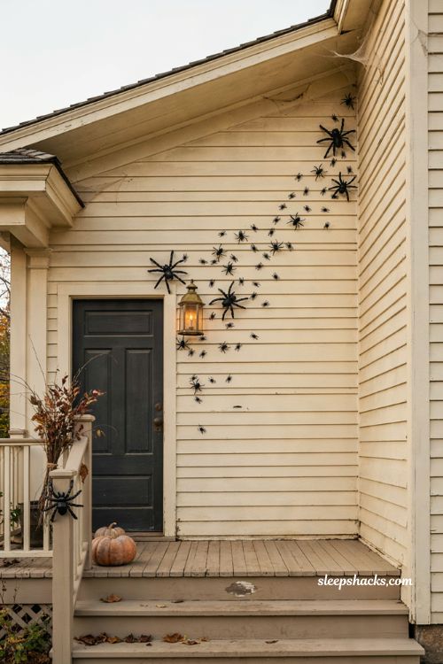 Halloween front porch with a trail of black spiders climbing light-colored house siding in an uneven diagonal path, oversized and small spiders mixed together, a few branching sideways, cozy cottage architecture, subtle pumpkins and warm lanterns, dramatic spooky facade, simple reusable Halloween decor, photorealistic editorial photography
