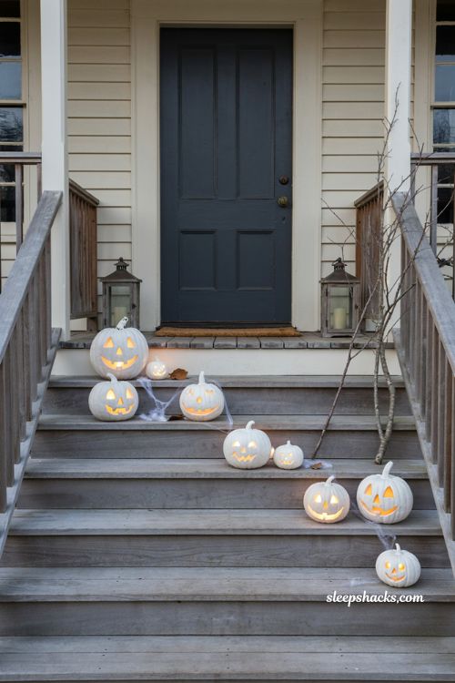 Monochrome Halloween front porch decorated with clusters of white pre-lit jack-o'-lanterns in varying sizes along the steps and porch floor, glowing softly at dusk, faux spiderwebs draped over the pumpkins, elegant cottage entryway, refined white and ivory Halloween decor, cozy spooky atmosphere, photorealistic editorial photography