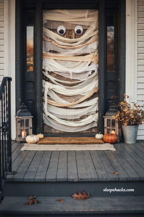 Mummified Halloween front door wrapped in tattered white gauze and crepe paper with oversized googly eyes, dark wood door contrasting with aged white fabric, cozy autumn porch, playful DIY Halloween decoration, small pumpkins and warm lanterns, charming spooky home entrance, photorealistic editorial photography