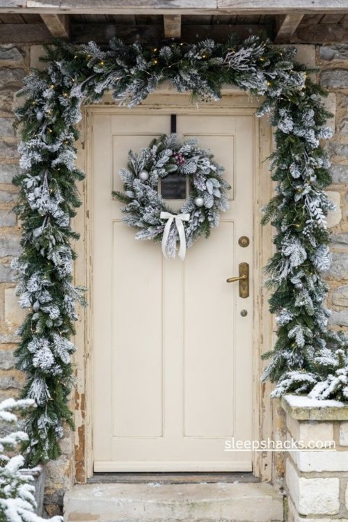 Flocked Christmas wreath and matching snowy garland with white ribbon and warm lights around the front door