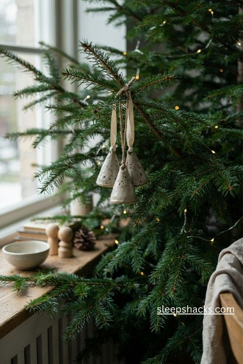 A medium-wide editorial interior photograph of an elegantly decorated Christmas tree in a warm, lived-in living room, showcasing a small collection of white ceramic bells with dipped gold metallic edges hanging near the front of the tree. The scene feels like a quiet December afternoon in a home where holiday decorations are chosen with care, blending the delicate beauty of handmade keepsakes with the comforting familiarity of traditional Christmas rituals. Timeless European cottage style with understated vintage elegance and subtle modern touches, featuring natural greenery, aged wooden furniture, soft textiles, and thoughtfully collected seasonal ornaments. A refined palette of warm ivory, creamy white, deep forest green, muted champagne gold, and natural honey-toned wood, with gentle variations in the metallic finish that keep the arrangement from feeling overly polished. Small white ceramic bells feature smooth glazed bodies and clean rounded silhouettes, each dipped halfway in reflective antique gold with a naturally uneven boundary that reveals the handmade character of the finish. Realistic gold clappers are visible inside a few bells, while short strands of soft cream velvet ribbon loop through the hanging openings. Several bells in slightly different sizes hang individually at varied heights from sturdy evergreen branches, their gold edges catching the light without overpowering the soft white ceramic. A few delicate porcelain stars, natural pinecones, and restrained gold accents appear farther back among the branches, while a weathered wooden console and loosely folded ivory linen add a subtle sense of home beyond the tree. The ceramic surfaces show gentle highlights, the metallic edges reflect warm surroundings, and the small clappers add a visual suggestion of a bright, delicate ringing sound whenever the branches move. Soft winter daylight enters through a nearby window and mingles with warm white Christmas lights, creating luminous reflections along the gold-dipped edges and natural shadows between the dark green needles. The mood is quietly festive, graceful, and nostalgic, like rediscovering beloved bells that have rung through many Christmas mornings, combining boutique-like elegance with the intimacy of family traditions. Soft colors that change the mood instantly, old textures that add warmth and depth, simple details that create lasting charm. Sophisticated but authentic editorial composition, realistic ceramic glaze and metallic reflections, intricate bell details, natural evergreen textures, balanced visual spacing, every ornament and surface sharply defined, rich material contrast, no excessive glitter, no artificial showroom styling, no people, no text, no watermarks, no blur, no bokeh, all surfaces in sharp focus, photorealistic, 4K resolution, editorial interior photography, Pinterest-worthy