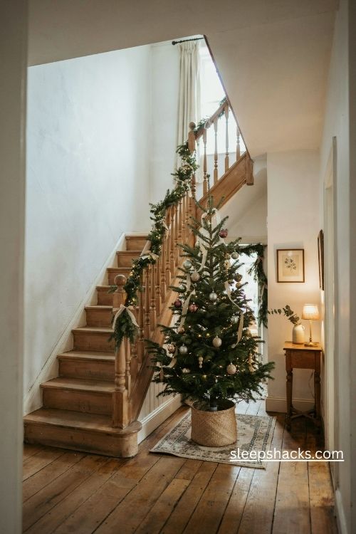 slim christmas tree on a staircase landing with glowing lights, evergreen banister garland, and cozy farmhouse decor